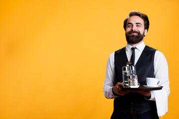Smiling butler poses with platter in hand, standing against yellow background while he works at...