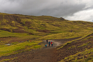 Obraz premium Tourists hike through mossy and wetland terrain of Reykjadalur Valley, Iceland. Valley is known for its mudpots, soda springs and geothermal activity.