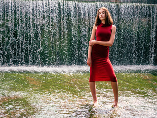 Young teenager girl in stylish red dress in unique location in water and water fall in the background. Lady in high fashion outfit for prom or special event in beautiful nature scene.