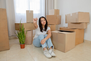 Portrait young woman sitting on the floor surrounded by moving boxes - young happy Hispanic woman...