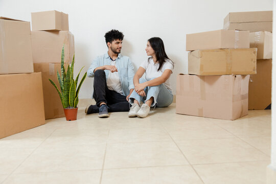 Portrait of happy couple moving into their new home - Hispanic couple sitting on the floor surrounded by moving house in their apartment