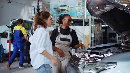 Certified repairman in auto repair shop showing customer what needs to be changed on her car for it to properly work. Garage expert inspecting vehicle placed on overhead lift, talking with woman
