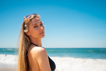 Blond woman on beach. Female with long hair and sunglasses on her head looking to camera while standing on sandy beach. Portrait of attractive girl in black swimwear with azure water and blue sky on