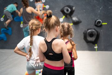 Three girls in sport clothes on climbing session. Back view of kids watching a coach in rock...