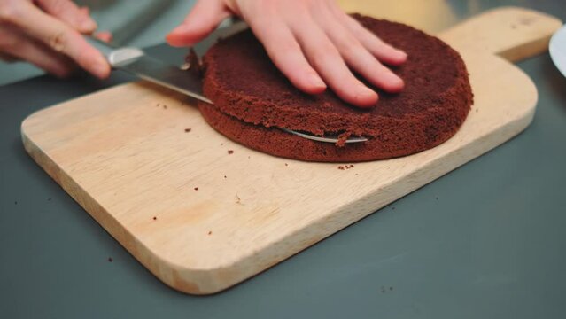 A woman cuts a chocolate biscuit cake with a knife into two thin parts. Middle shot of the kitchen table. Cooking in the kitchen