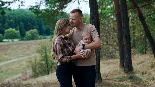 Happy young parents with their firstborn baby. Caucasian couple stand in nature with their newborn. Man kisses his wife on the forehead.
