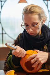 A modern blonde woman in military uniform is carving spooky pumpkins with a knife for Halloween...