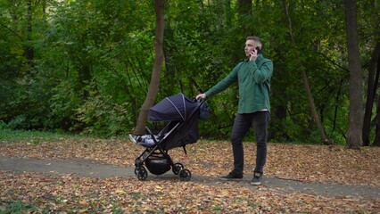 Obraz premium A young dad is talking on the phone and rocking his child in a stroller. A man speaks on the phone in the autumn forest.
