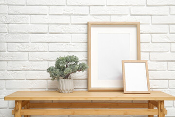 Wooden table with tree in pot and blank frames near white brick wall