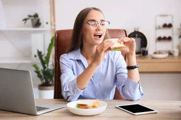Fotobehang Beautiful young businesswoman eating tasty sandwich at her workplace in office © Pixel-Shot