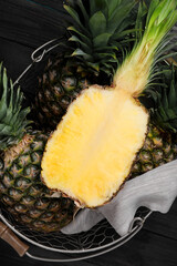 Whole and cut ripe pineapples in metal basket on black wooden table, top view