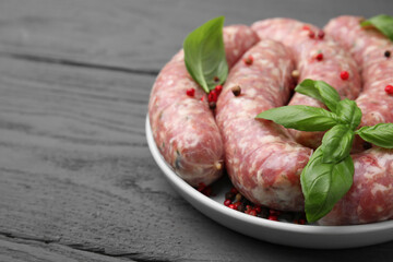 Raw homemade sausages, basil leaves and peppercorns on grey wooden table, closeup. space for text