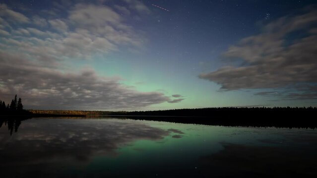 Evening time lapse video of a starry night with clouds moving over a very calm lake.  Green Aurora with patches of red light up the sky and reflect in the still water.
