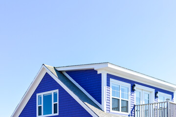 The roof of a vibrant purple-colored cape cod beach house. The trim on the peaked roof is white. There are multiple glass windows in the building with a patio door and a railed patio. The sky is blue.