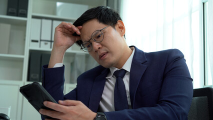 serious asian business man in suit using computer on desk disturb by mobile phone he is upset and busy in office hands on forehead
