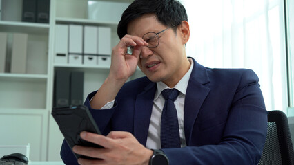 serious asian business man in suit using computer on desk suffering eye strain pain took off...