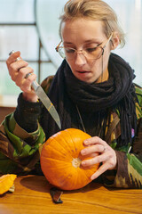A modern blonde woman in military uniform is carving spooky pumpkins with a knife for Halloween...
