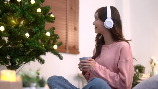 Young Asian woman wearing headphone to listening to music and holding cup drinking coffee by christmas tree at home, winter holiday concept.