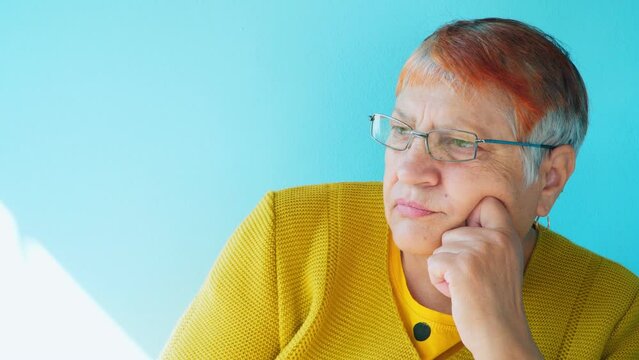 An elderly 70-year-old woman with glasses and gray hair looks thoughtfully to the side. Elderly life concept.