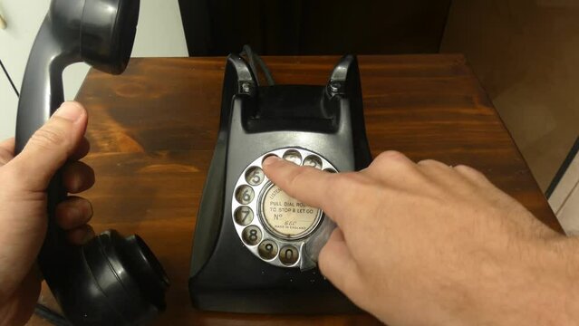 4k clip of an classic old black rotary dial phone. A hand picks up the headset and starts to dial using the rotary dial. Putting the finger in the desired number and turning the dial.