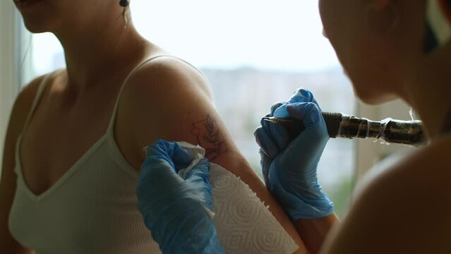 Tattoo artist makes floral tattoo. Tattooist in gloves working with professional tool, tattoo master wipes protruding blood from shoulder after tattooing. Close up