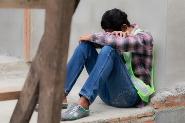 Hopeless builder worker sitting in construction site bend head down in his knee, stressed Asian...