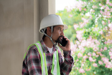 Asian or Indian laborers man wearing safety reflected vest sitting at townhouse construction site or resort talking to a contractor using mobile phone, foreman relaxing communication with colleagues
