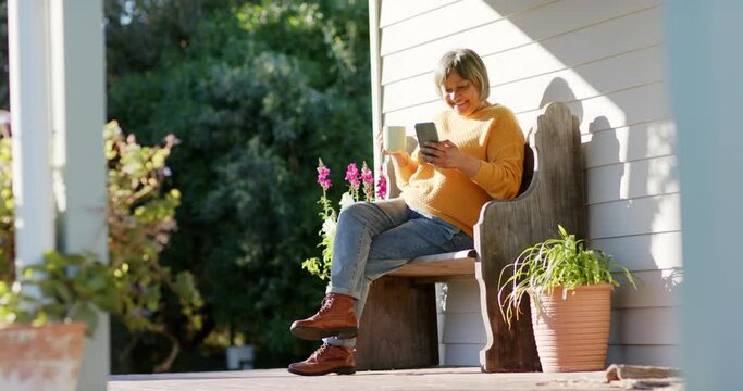 Happy senior biracial woman using smartphone and drinking coffee on terrace, slow motion, copy space