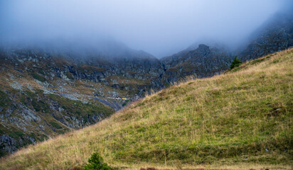 Wonderful view of rocky mountains partially covered by fog and a slope with a meadow.