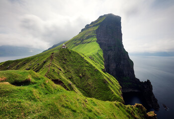  lighthouse sitting on top of a grassy hill close to a rocky black cliff looks over the tranquil sea on cloudy day. Breathtaking aerial view of a small green islet in Faroe Islands. - obrazy, fototapety, plakaty