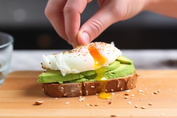 hand spreading avocado on toast, placing poached egg over top - obrazy, fototapety, plakaty