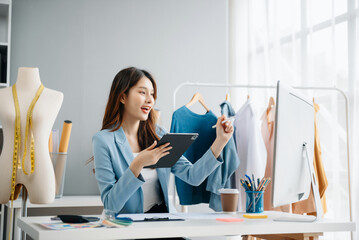 Fashion designer woman talking smart phone and using laptop with digital tablet computer in modern studio the clothes hanging on the racks