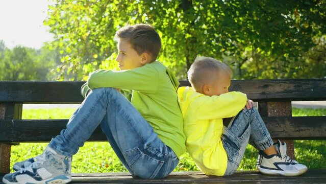Younger and older offended brothers sit back to back on a bench after an argument. Siblings misunderstanding, ignoring each other after quarrel. No compromising, not talking. Rivalry concept