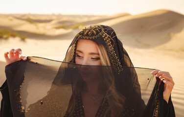 Desert adventure. Young arabian Woman posing in traditional Emirati dress abaya in sanddunes of UAE desert at sunset. The Dubai Desert Conservation Reserve, United Arab Emirates. - obrazy, fototapety, plakaty
