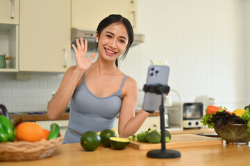 Obraz na płótnie Canvas Fit young asian girl drinking green smoothie and recording her blog about healthy food on smartphone at the kitchen