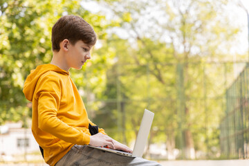 Thoughtful teenager boy resting. Holding and using a laptop for networking on a sunny day, outdoors. - obrazy, fototapety, plakaty