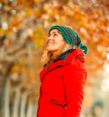 Young happy cheerful adult woman posing outdoor and enjoying leisure activity wearing a red warm jacket. Autumn colors and trees in background. Cute middle age female people at the park smiling