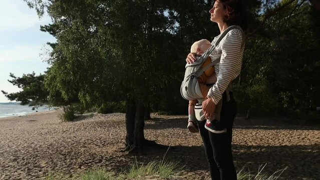 a 30-year-old woman with a child in an ergo-backpack sling stands on the beach against the backdrop of pine forest and looks at the sunset, enjoying the peace and tranquility