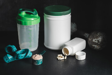 Chocolate whey protein powder in measuring spoon, white capsules of amino acids and vitamins, measuring tape, old rusty dumbbell, plastic shaker on dark background. bodybuilding food supplements