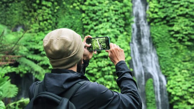 Adult Man Traveling in Green Nature Takes Phone Photo of Forest Waterfall. Lifestyle of Young Unrecognizable Person at Trip Destination of Jungle Water Fall. Concept of Traveler Making Journey Videos