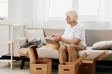 elderly woman sits on a sofa at home with boxes. collecting things with memories albums with photos...