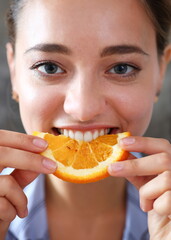 Woman portrait holds in her hand a slice of chopped orange eats her for breakfast with her mouth in the kitchen. Concept of a healthy diet.