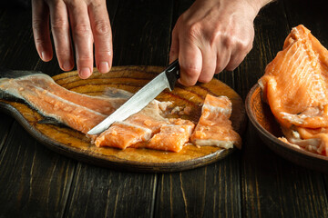 Close-up of a chef's hands with knives while cutting or cleaning fresh red fish on a kitchen cutting board before preparing a fish dish for lunch.