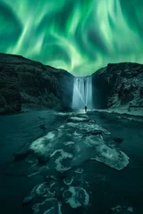 Foto auf Glas Aurora Borealis dancing over the frozen Skógafoss Waterfall in winter. A man with a lantern is watching dance across night skies the Aurora Polaris next to the famous Skógafoss waterfall, Iceland © Revive Photo Media