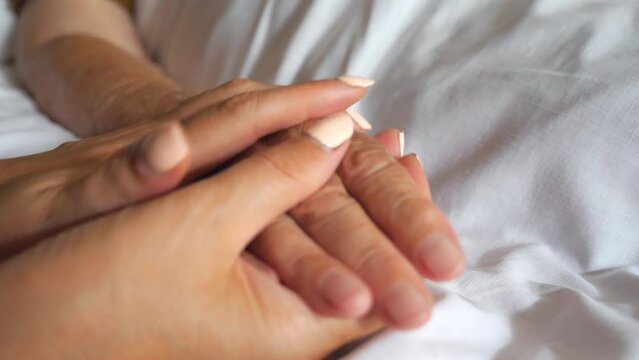 Young woman gentle stroking hand of mother taking care her. Daughter comforting wrinkled arm of elderly mom lying at bed in hospital. Girl showing love to parent. Detail view Slow motion Dolly shot