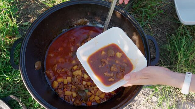 Cook pours red pepper soup with tomato and meat in a disposable plastic bowl. Close-up top view of huge cauldron with bograch or borsch in outdoor and woman hands hold kitchen ladle. Camping season