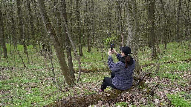 Back view of young woman in baseball cap filming video or taking photo pictures on his mobile cell phone during his adventure in the forest. Blogger girl in a woodland shooting content on smartphone