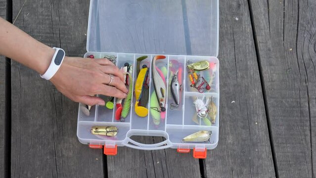Woman hand open large fisherman's tackle box takes one bait and closes the box. Case fully stocked with lures and gear for fishing. Fish lures and accessories for spinning. Kit of fishing lure closeup