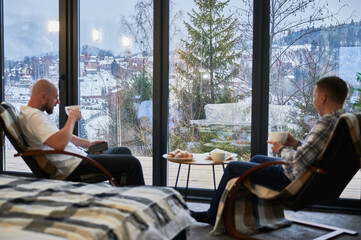 Focus on winter mountain landscape through panoramic windows in contemporary barn house. Two young men sitting on chair, holding cup of tea, enjoying view of magical scenery on blurred foreground.