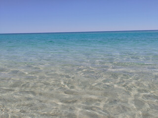 Une plage côtière tranquille avec des eaux cristallines, différentes nuances de bleu clair sous un ciel azur et un soleil éclatant.